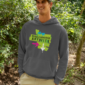 Smiling man outdoors in a forested area, wearing a gray hoodie with a bright Middlesbrough Art Week graphic on the front.